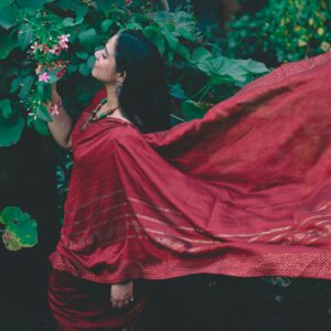 Woman in a flowing red saree stands among lush green foliage, embracing traditional Indian beauty.
